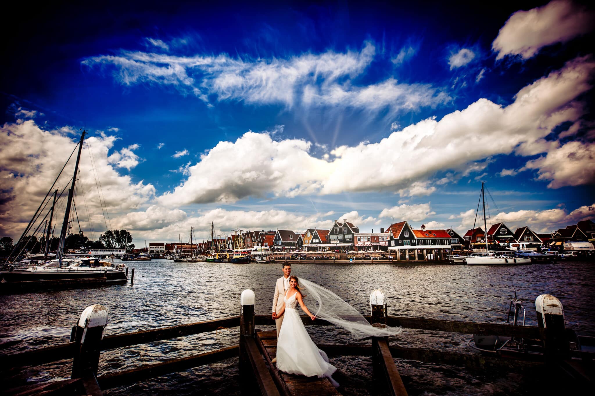 Het pasgetrouwde stel deelt een intiem moment op de Volendamse dijk, met een pittoresk uitzicht op de haven en de traditionele boten die rustig in het water dobberen.